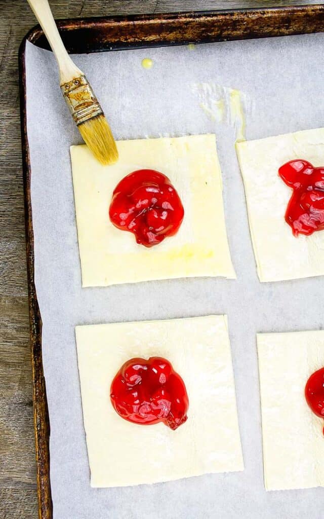 four squares of puff pastry with pie filling one with egg wash in the center on a baking sheet