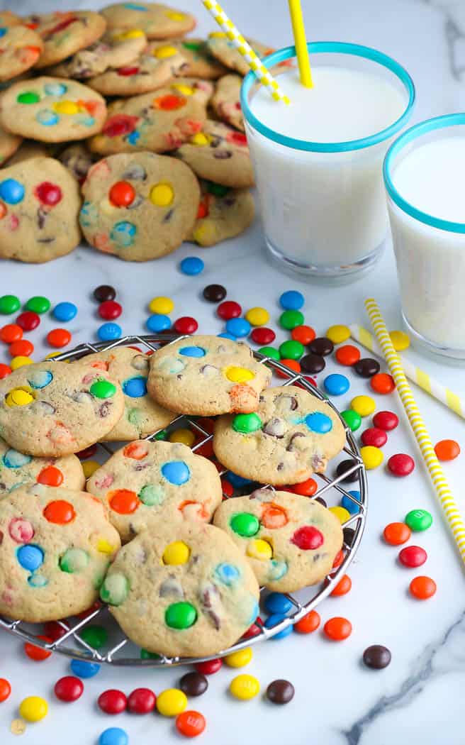 M&M Cookies on a wire rack next two two glasses of milk with yellow straws