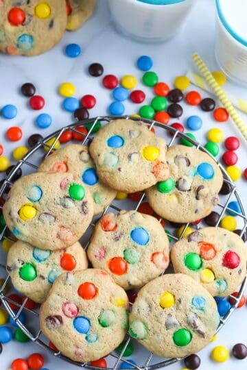 overhead picture of cookies with M and M candy on cooling rack with two glasses of milk and yellow straw