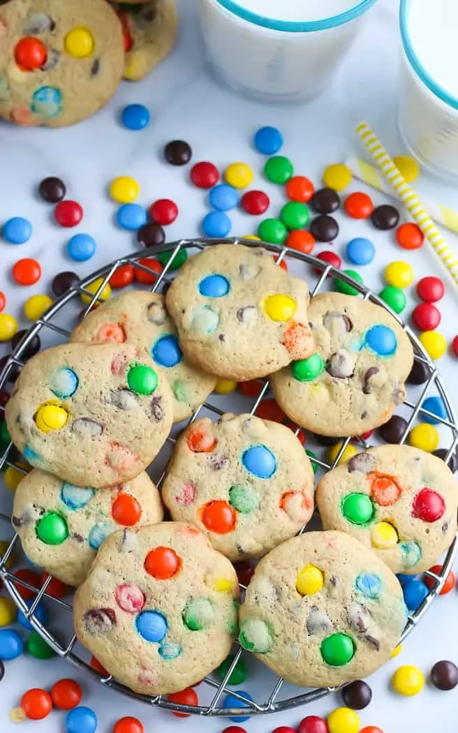 overhead picture of cookies with M and M candy on cooling rack with two glasses of milk and yellow straw