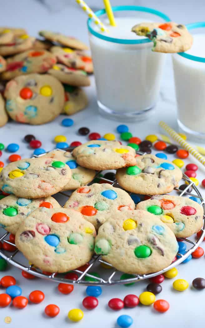 side view of M&M cookies on a cooling rack with glasses of milk in the background with a cookie balanced on them
