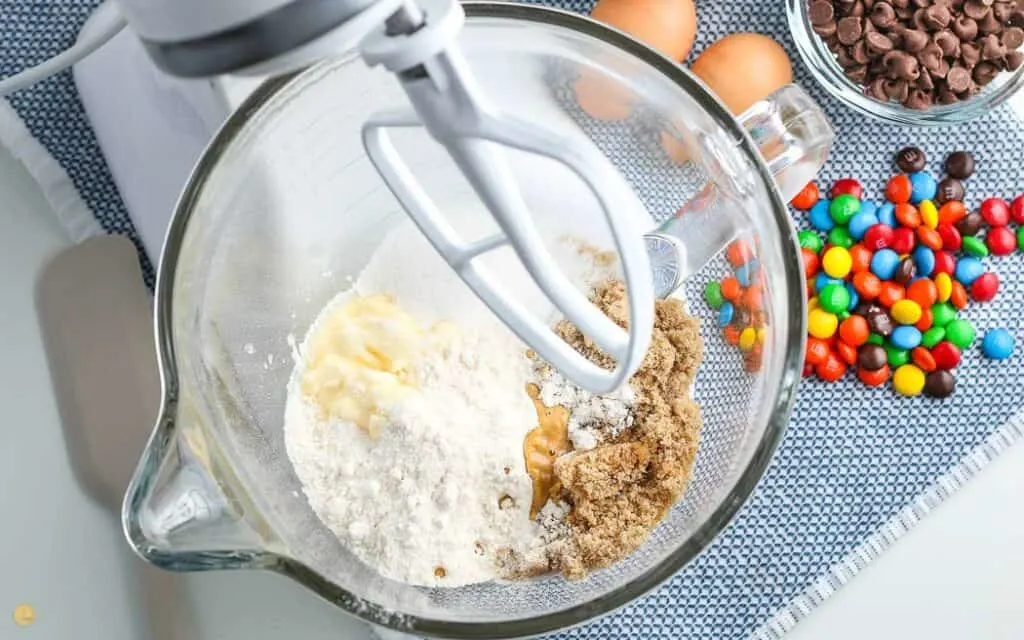 over head picture of stand mixer with cookie dough ingredients in the bowl on a blue placemat with a bowl of M&Ms next to it