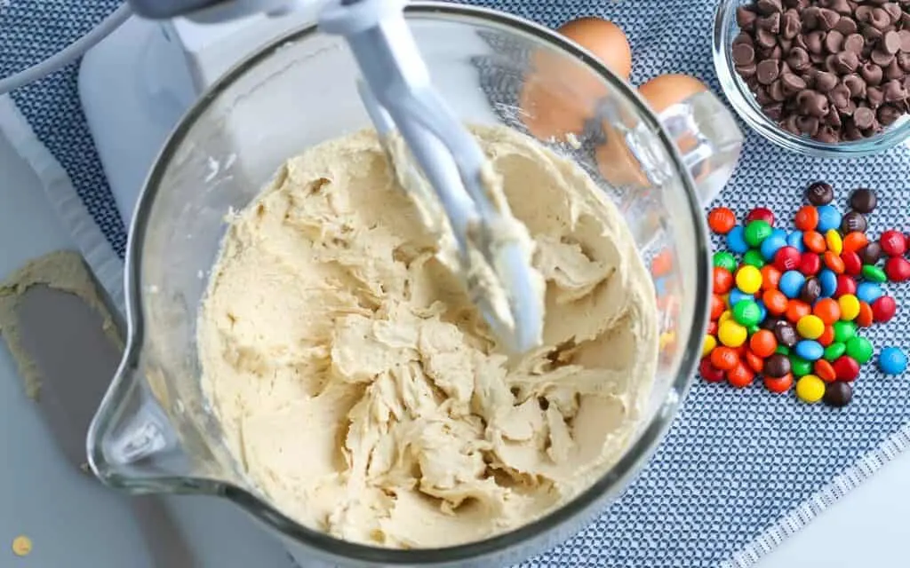 over head picture of stand mixer with mixed cookie dough ingredients in the bowl on a blue placemat with a bowl of M&Ms next to it
