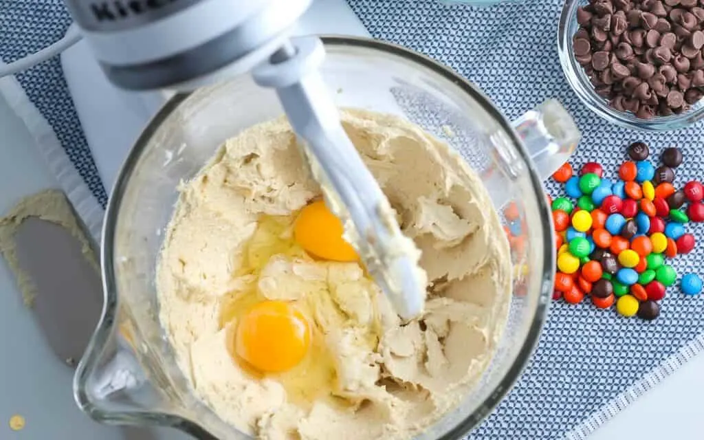 over head picture of stand mixer with cookie dough and eggs in the bowl on a blue placemat with a bowl of M&Ms next to it