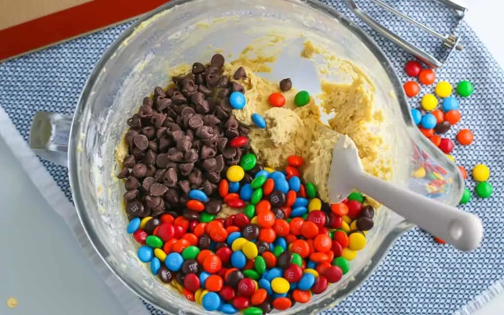 mixing bowl with cookie dough, M&M candies, and chocolate chips with a spatula on a blue placemat