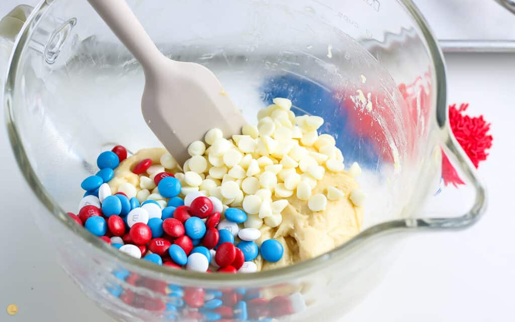 clear mixing bowl with cake batter, white chocolate chips, red white and blue m&ms, and a spatula