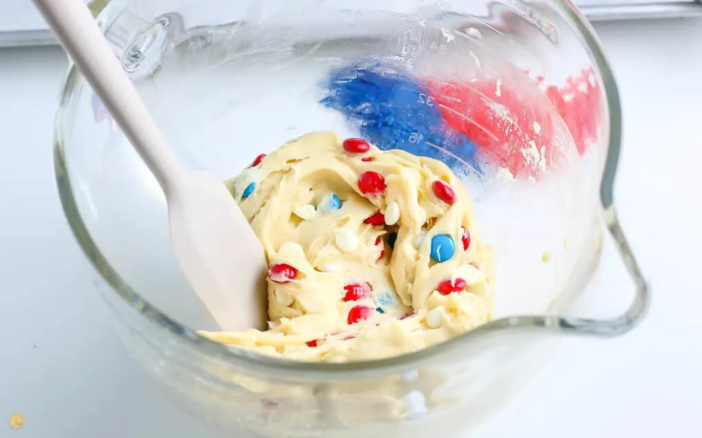 clear mixing bowl with cake batter, white chocolate chips, red white and blue m&ms all mixed up with a spatula