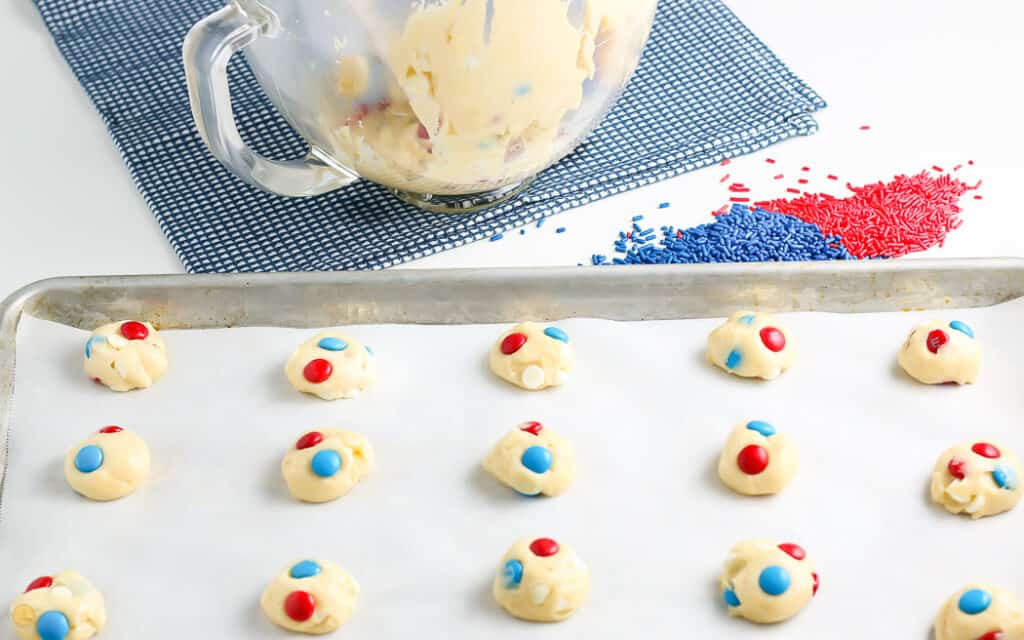 baking sheet with parchment paper and red white and blue m&m cookie dough balls with a mixing bowl in the background and two piles of sprinkles