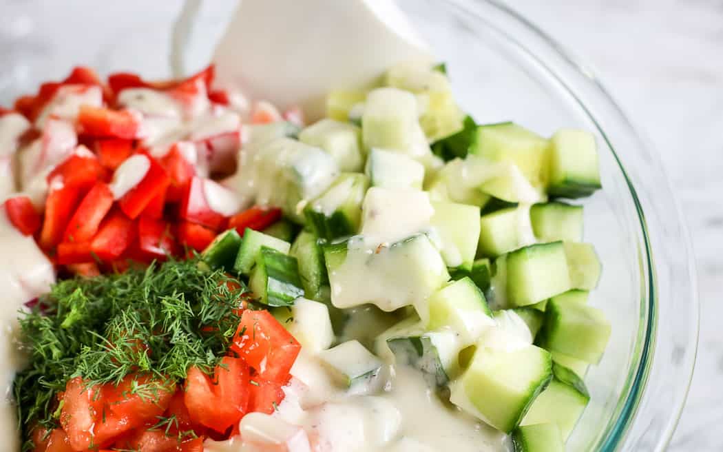 unmixed chopped tomatoes and cucumbers in a clear bowl with dressing poured on top with a white spatula