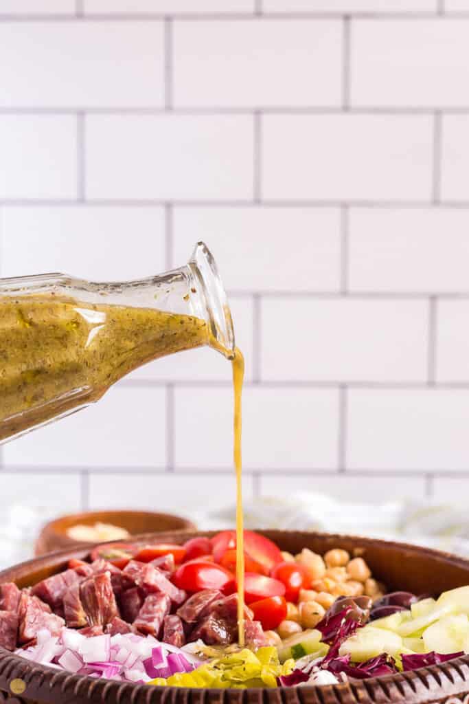 salad dressing being poured onto a salad in front of a white tile wall