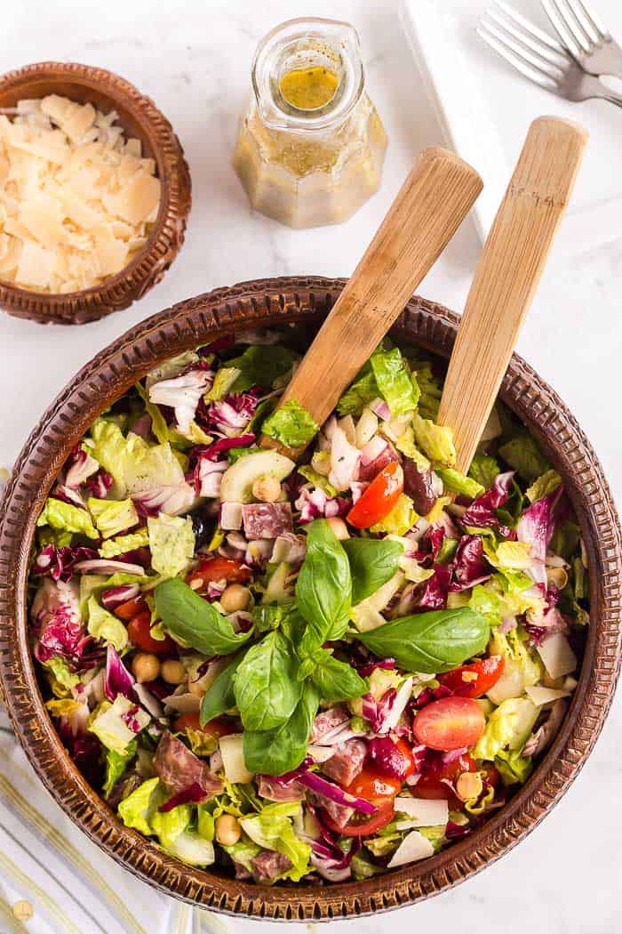 mixed Italian chopped salad in a wood bowl with wood serving spoons in it. Cheese and dressing off to the side