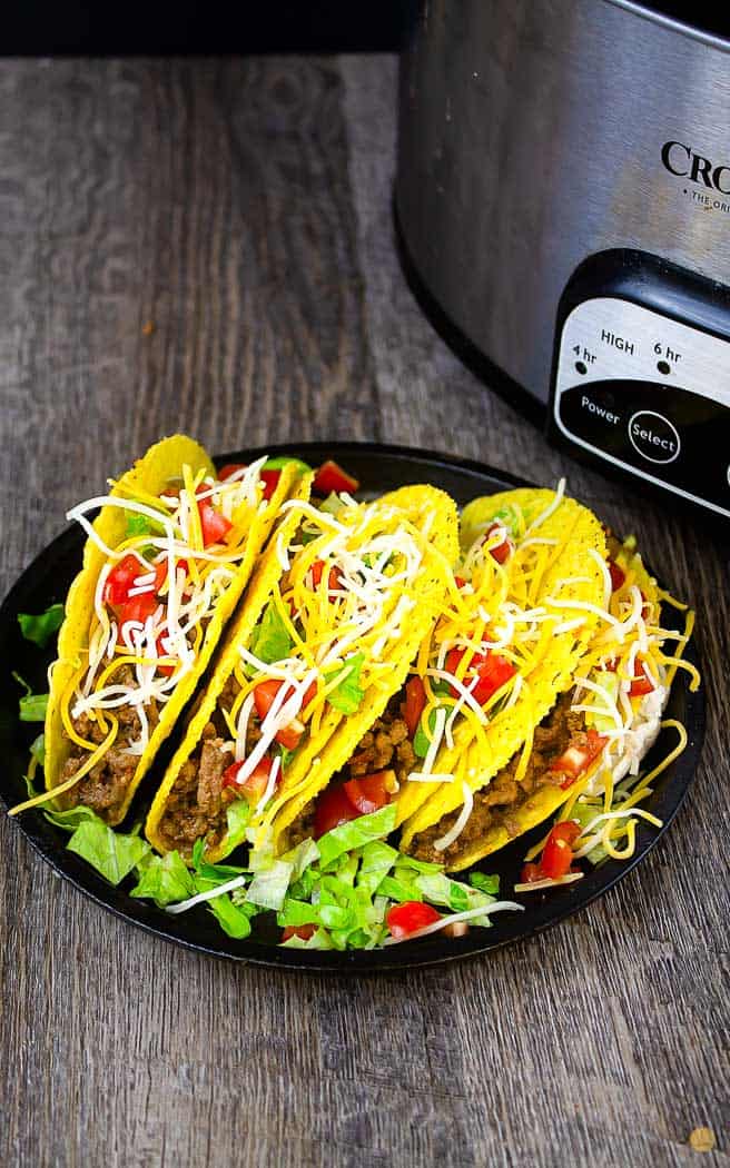 4 filled tacos on a black plate next to a crock pot