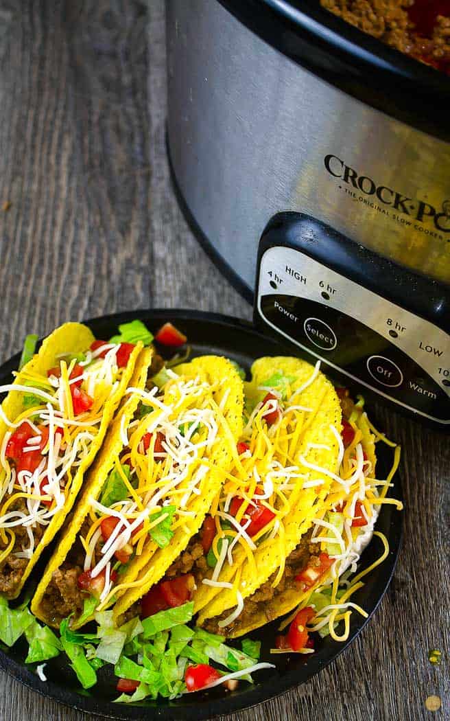 four dressed hard tacos on a black plate next to a crockpot