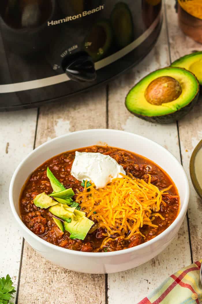 white bowl of chili topped with cheese, sour cream, and avocado