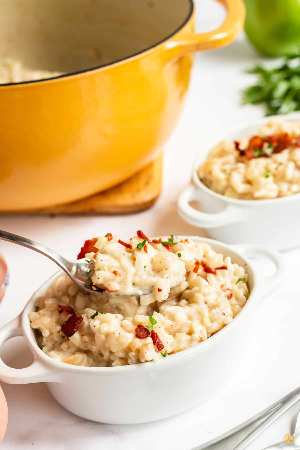 Risotto in a white bowl with bacon on top and a spoon in the bowl, on the table. A yellow pot is in the background as well as a 2n portion of mac and cheese.