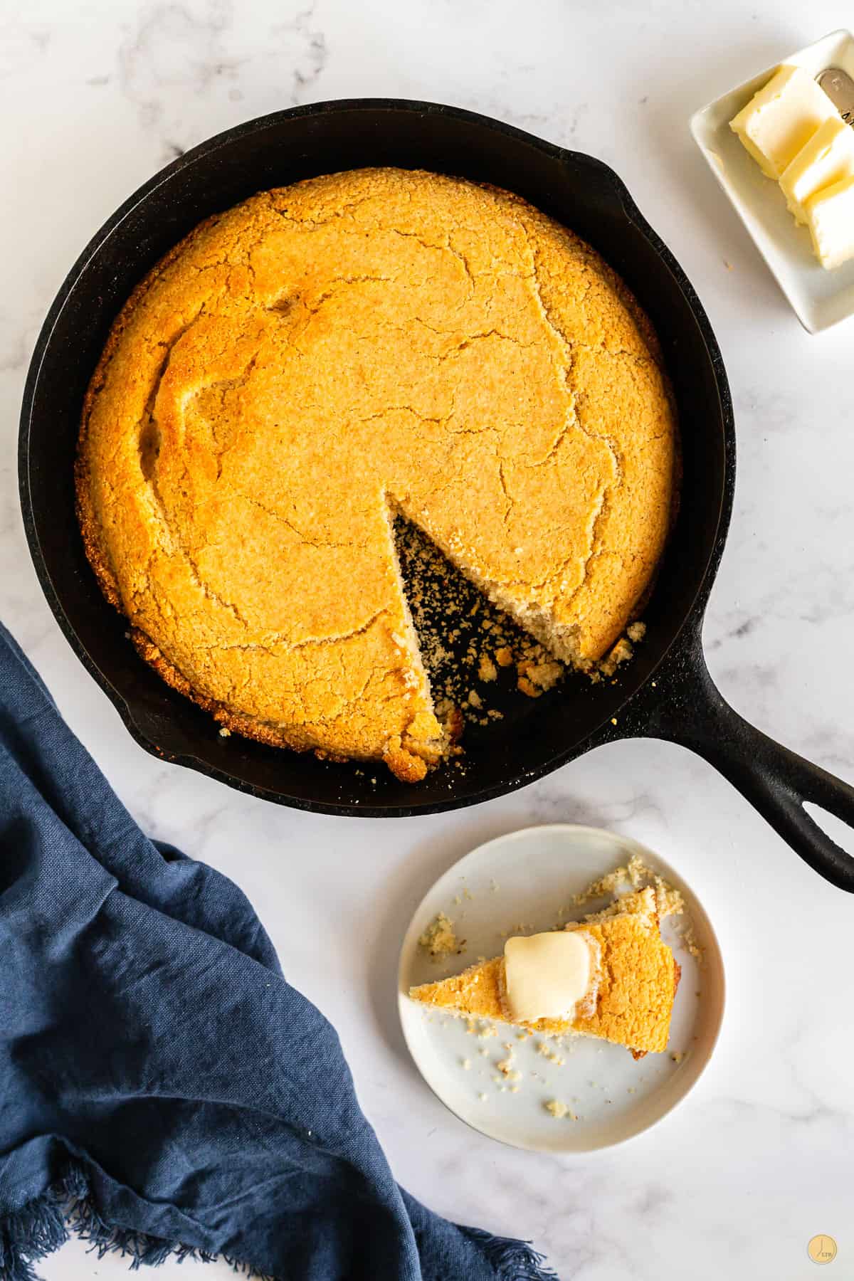 Top view of skillet with cornbread in it on a table, a slice cut out and is on a small white plate on the table next to it with butter on top.