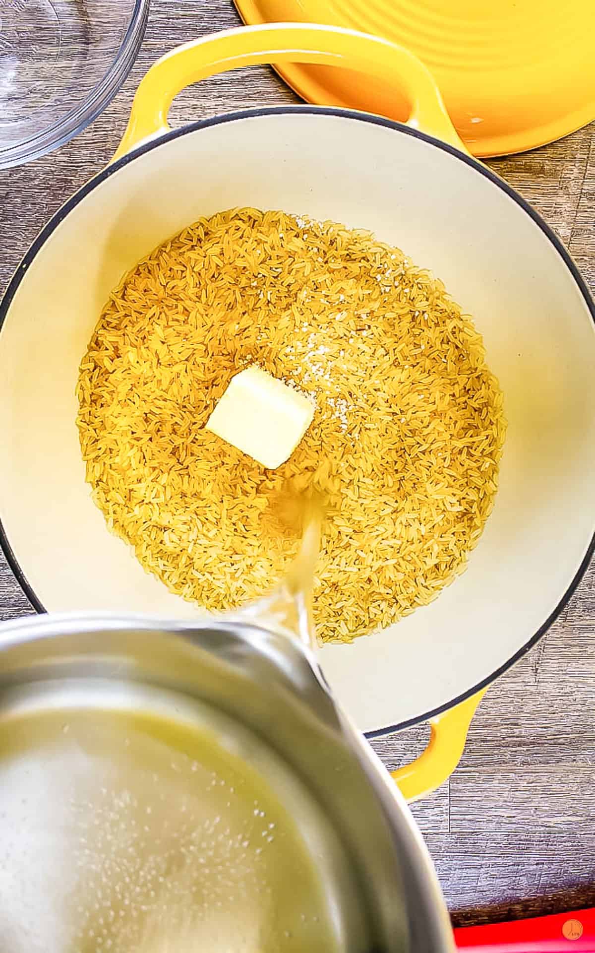 uncooked rice and butter in a dutch oven with water being poured into it.