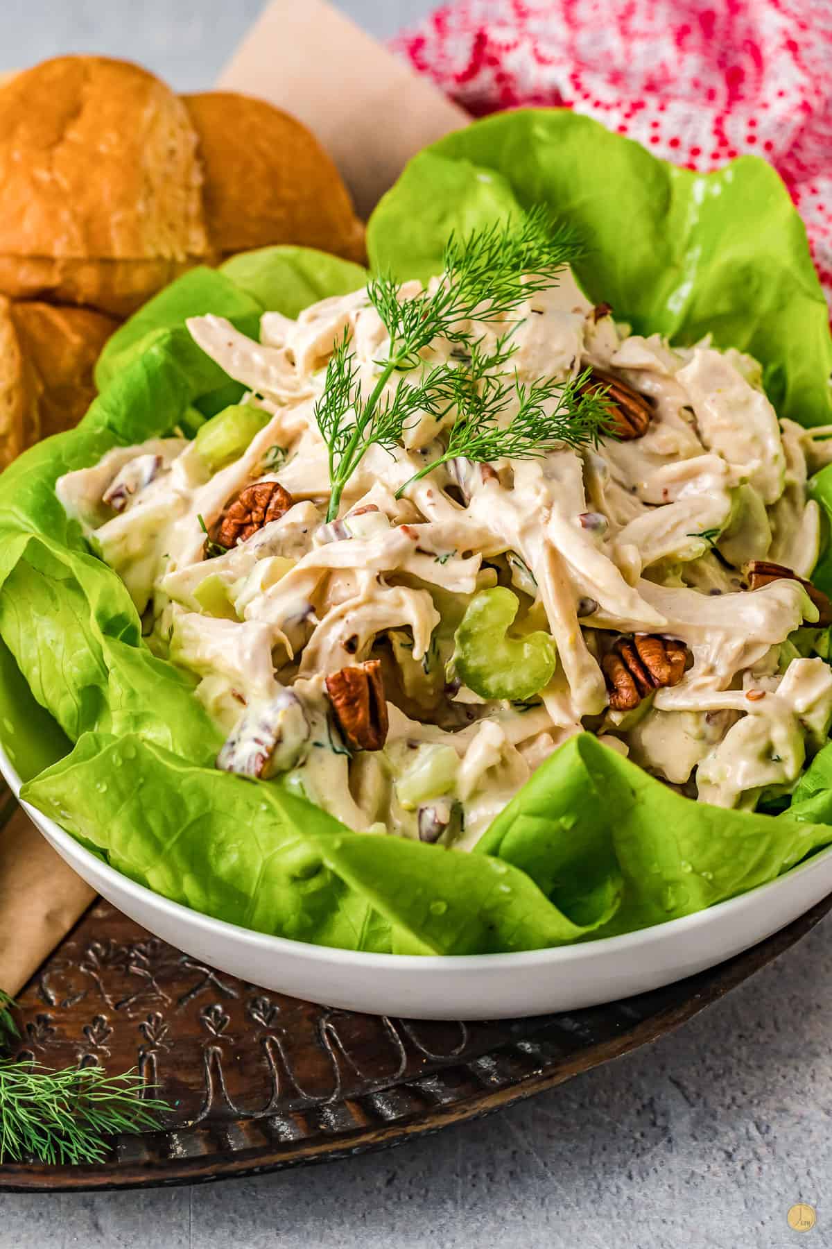 Creamy chicken salad with pecans served on a lettuce leaf, garnished with fresh herbs, in a white bowl on a table with rolls.