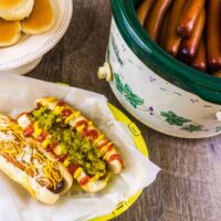 two dressed hot dogs in a paper basket next to a crockpot full of hot dogs