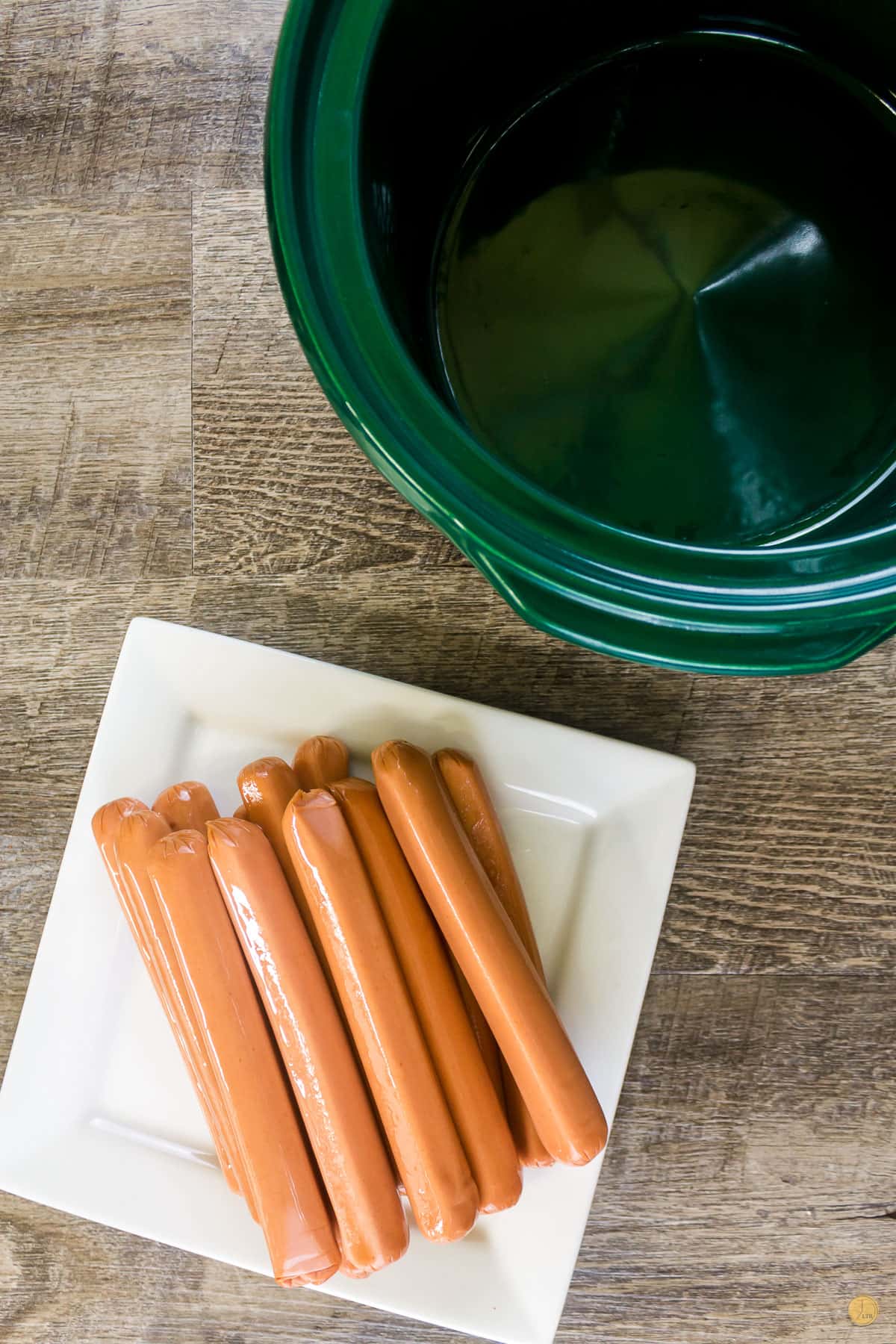 uncooked hotdogs on a white plate next to an empty green crock pot bowl insert