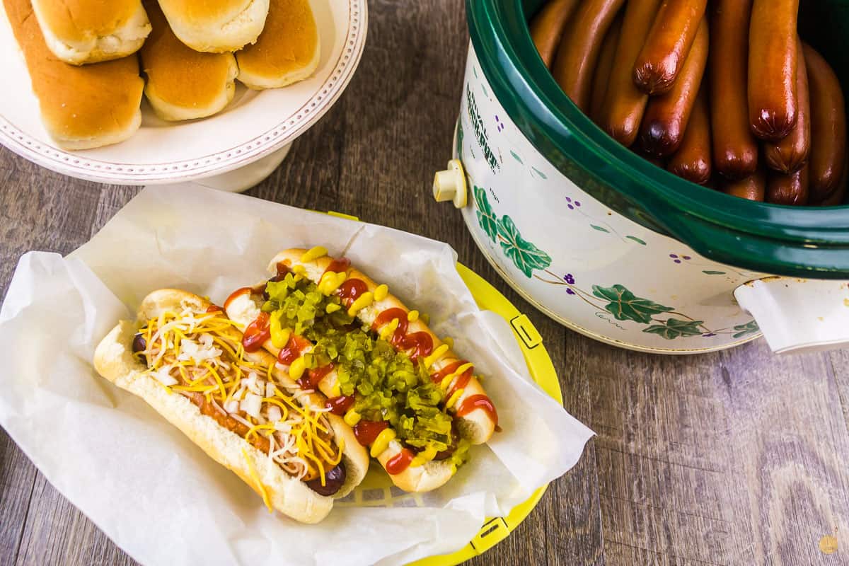 two dressed hot dogs in a paper basket next to a crockpot full of hot dogs