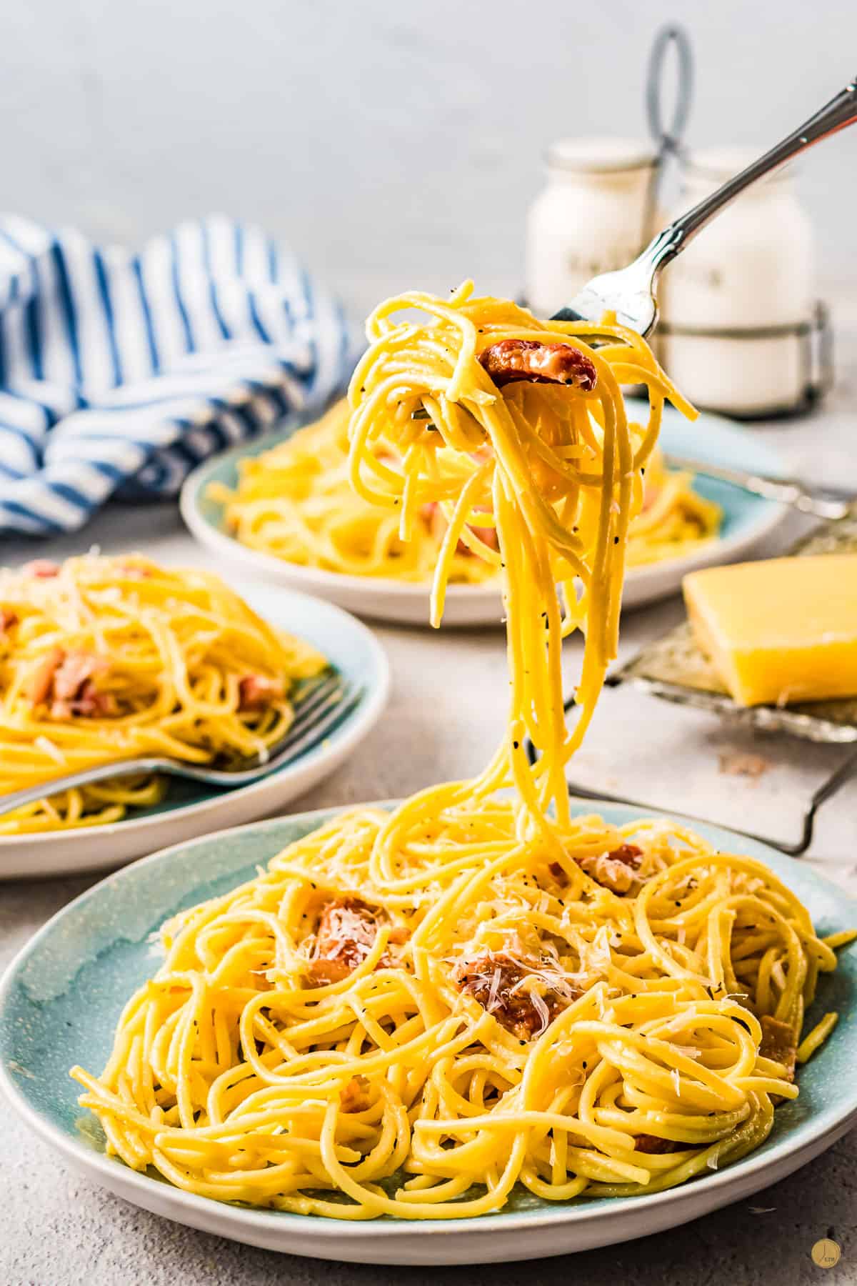Spaghetti Carbonara on a plate with a forkful of it being lifted up in mid-air. There are two other plates of spaghetti carbonara in the background, and it is all sitting on a table. 