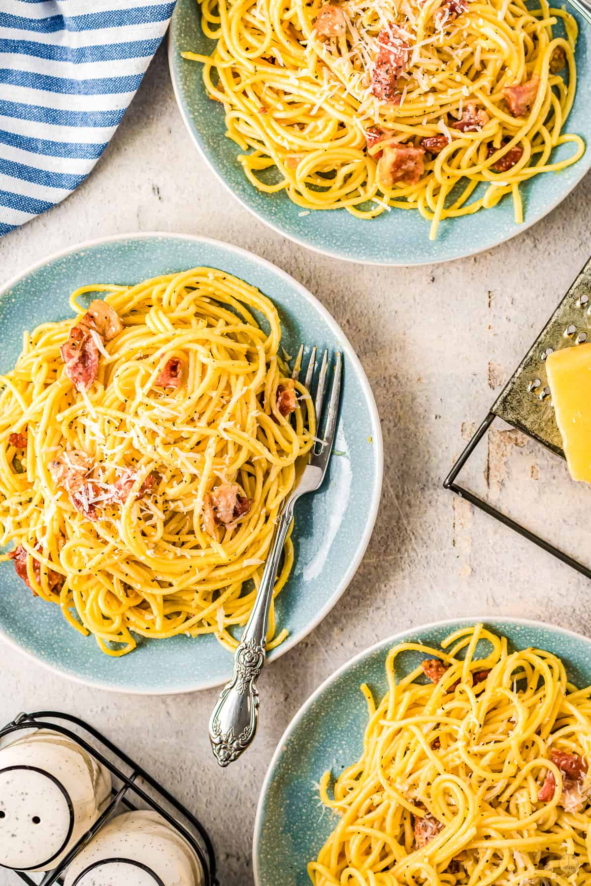 Spaghetti Carbonara on a blue plate . There are two other blue plates of spaghetti carbonara in the background, and it is all sitting on a table.