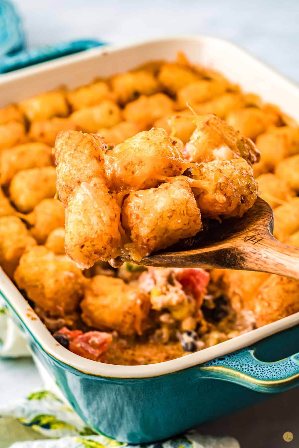 A blue and white dish filled with cowboy casserole with tater tots and cheese on top, with a wooden spoon lifting out a portion in mid-air. 