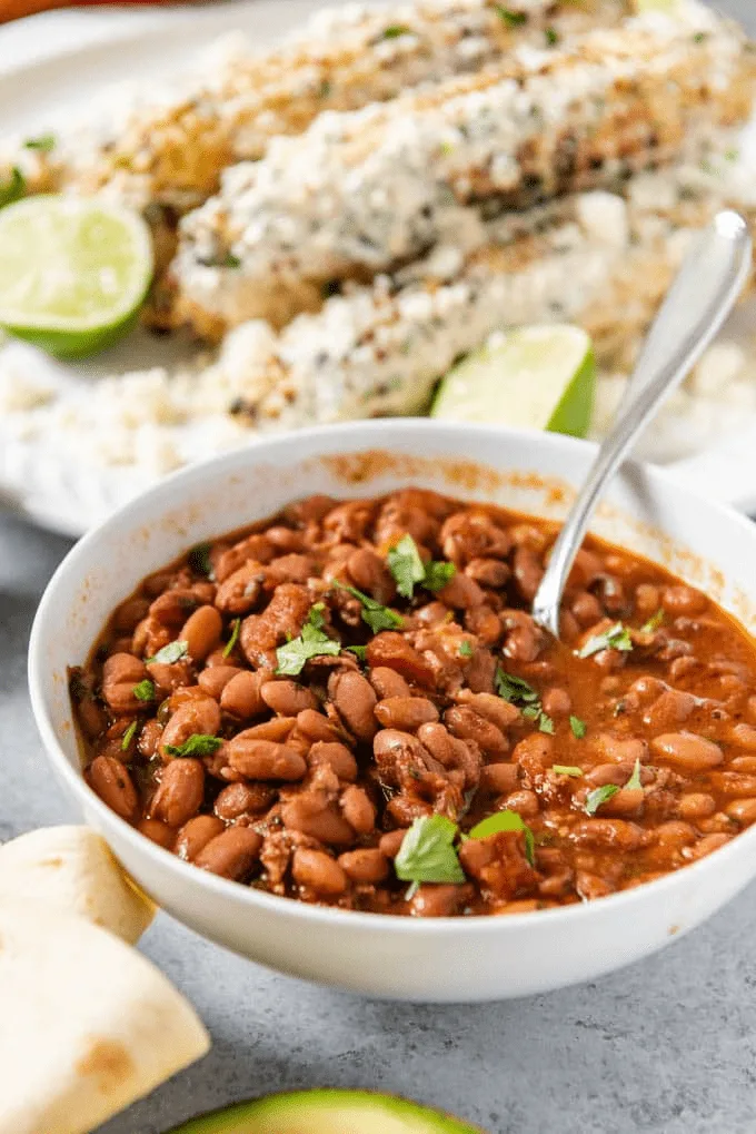 Charro Beans in a white bowl with a spoon in it and some grilled corn on the cob behind it, all on a table.