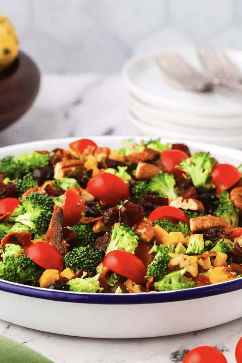 Broccoli salad with craisins, tomatoes and nuts in a white bowl on a table.