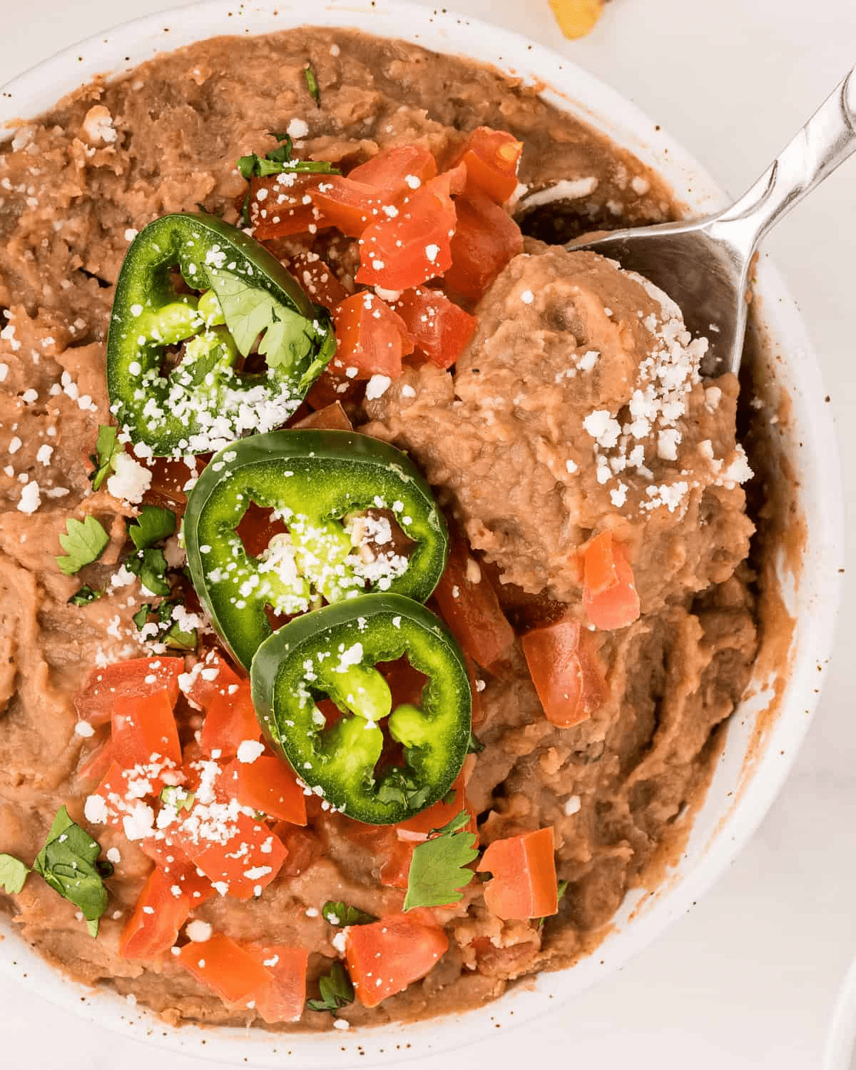 Refried beans in a bowl with pepper and jalapenos on top and a spoon inside the bowl.