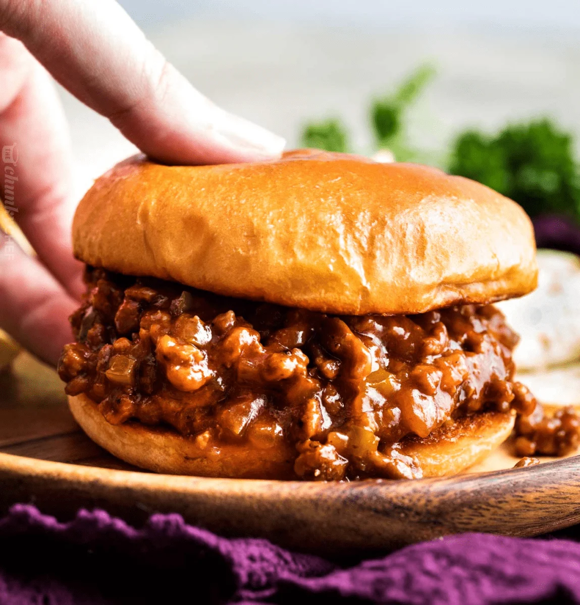 A hand reaches down to grab a bread bun filled with sloppy joe mince mix that is on a wooden plate. 