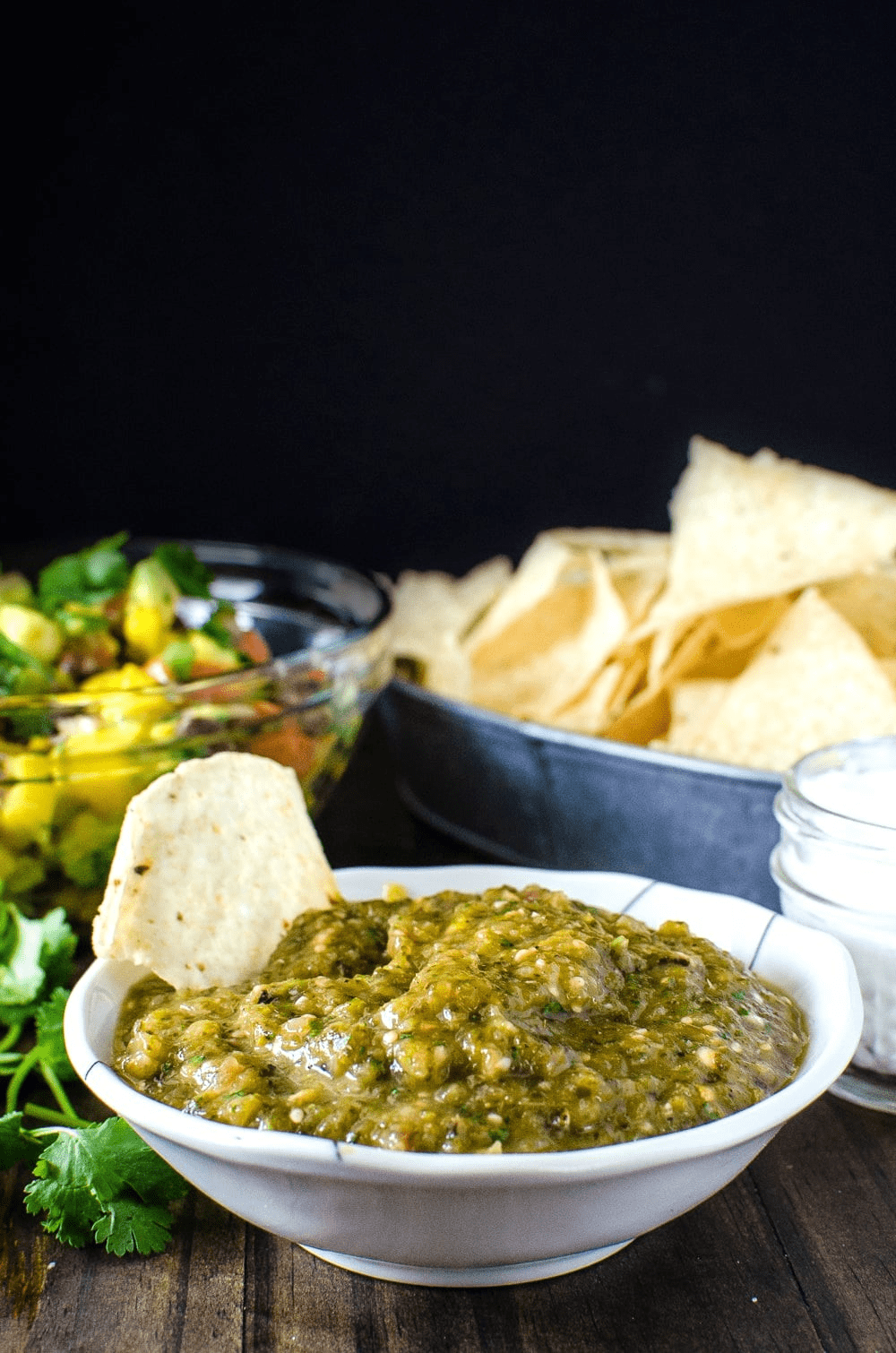 Green Salsa Dip "Roasted Tomatillo Salsa Verde" in a white bowl on a table with a bowl of chips and a bowl with salad in the background. 