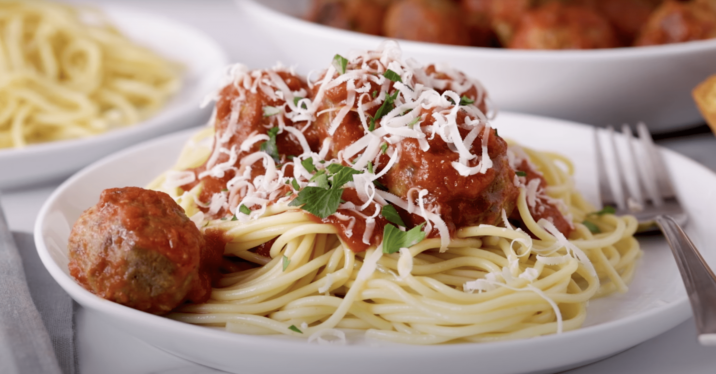 A white plate filled with meatballs lightly coated in a tomato sauce, on a bed of spaghetti, with herbs and parmesan cheese sprinkled on top. 