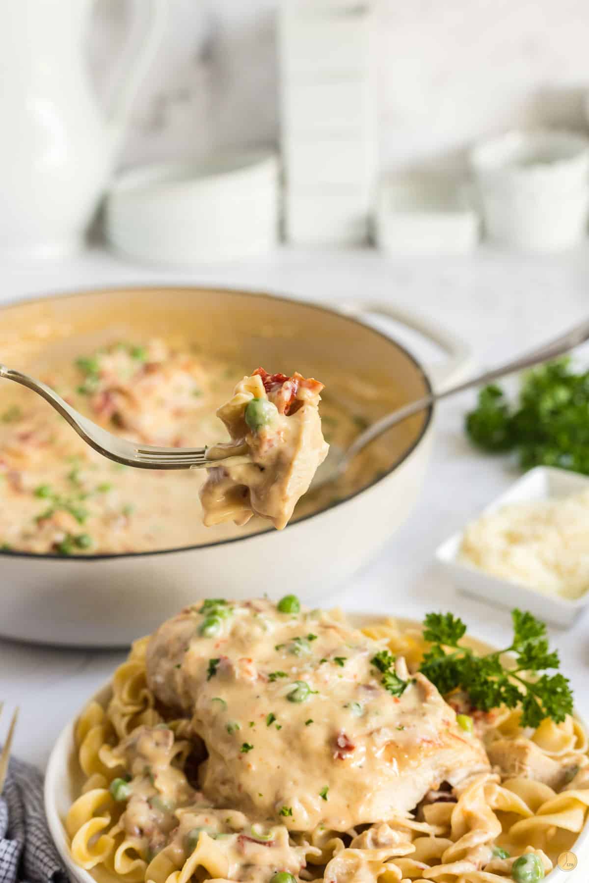 fork with bite of chicken over a plate of noodles with pan of creamy garlic chicken