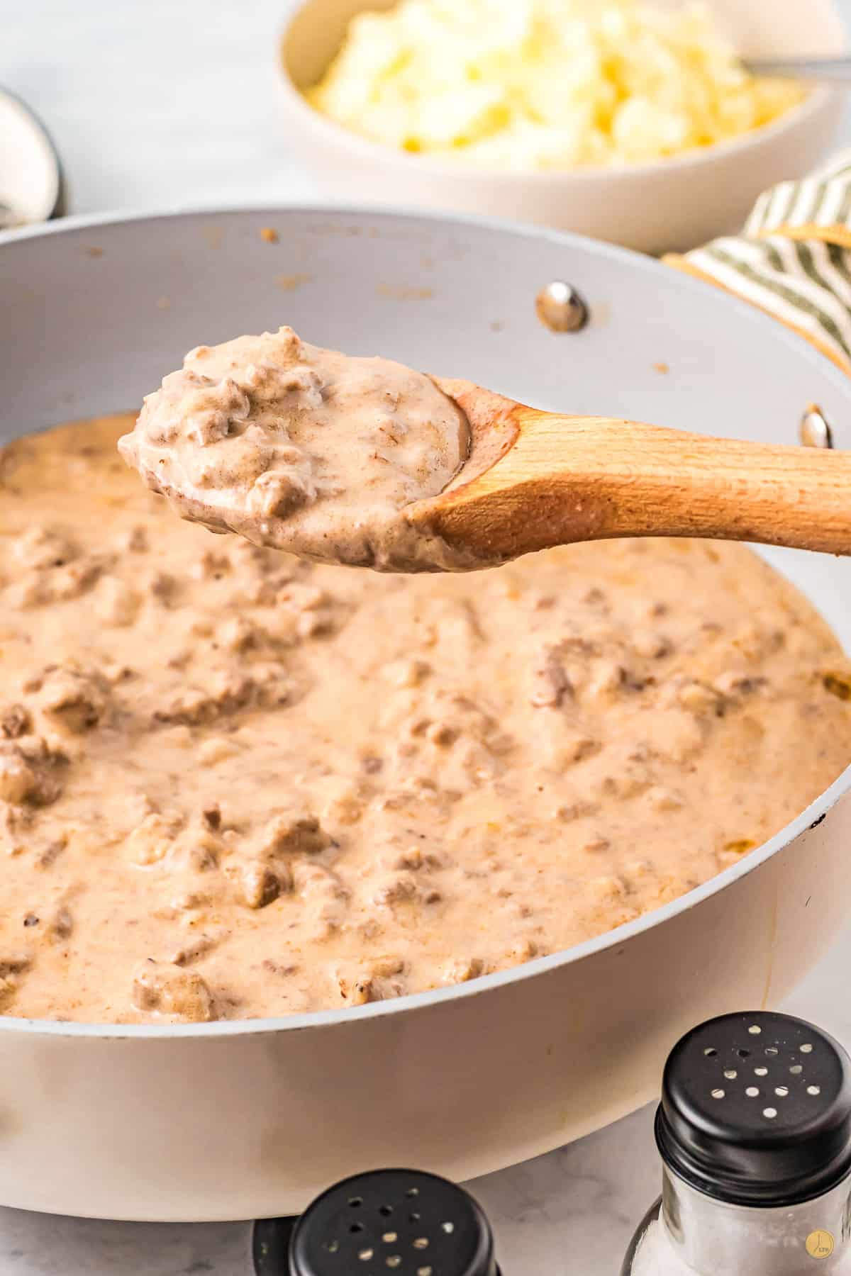 Close up of a large pan with hamburger gravy in it and a wooden spoon being held in mid-air filled with a spoonful of hamburger gravy. 
