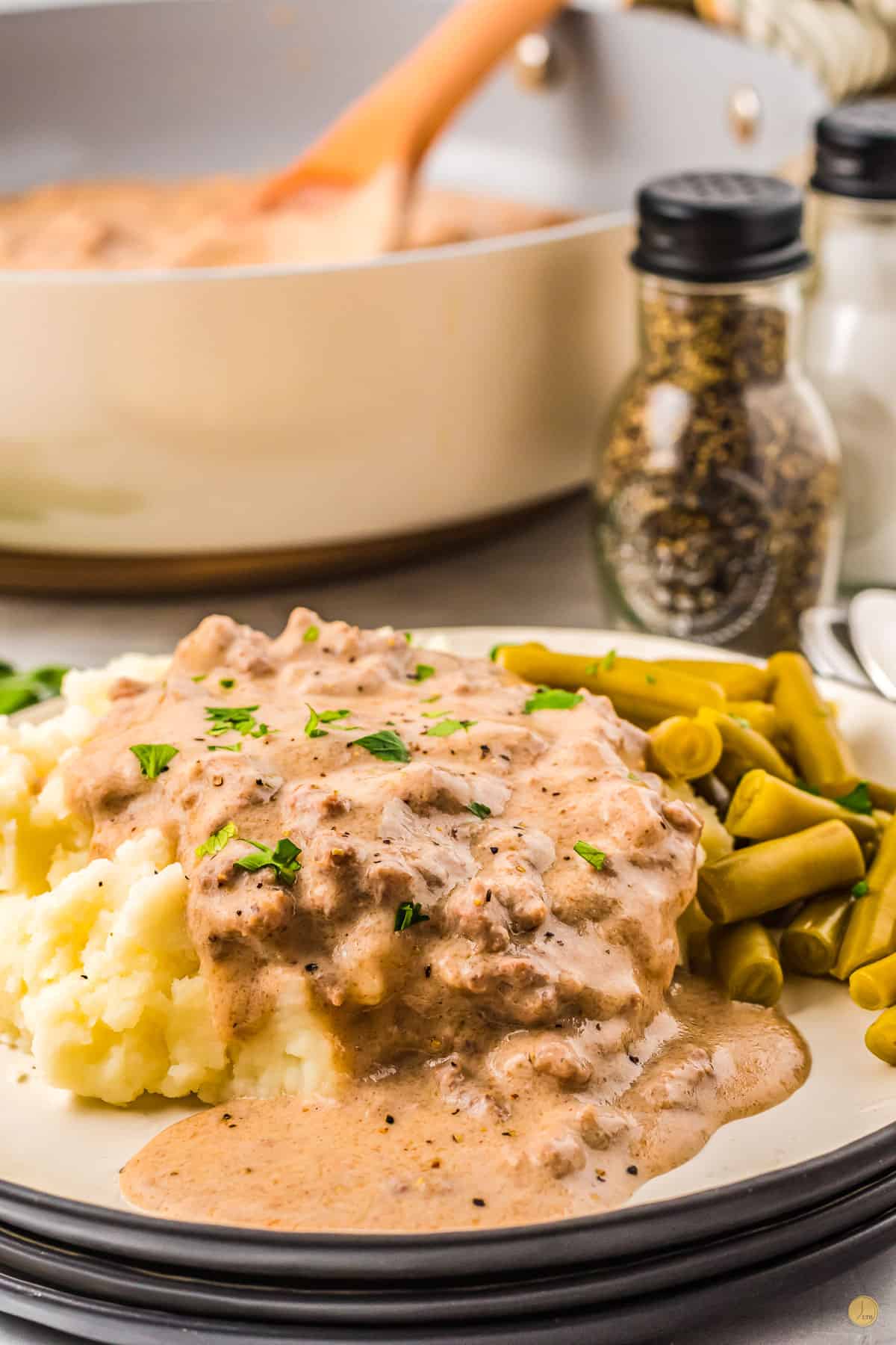 Close up of a white plate with a black rim filled with mashed potatoes topped with sausage gravy with a side of green beans. 