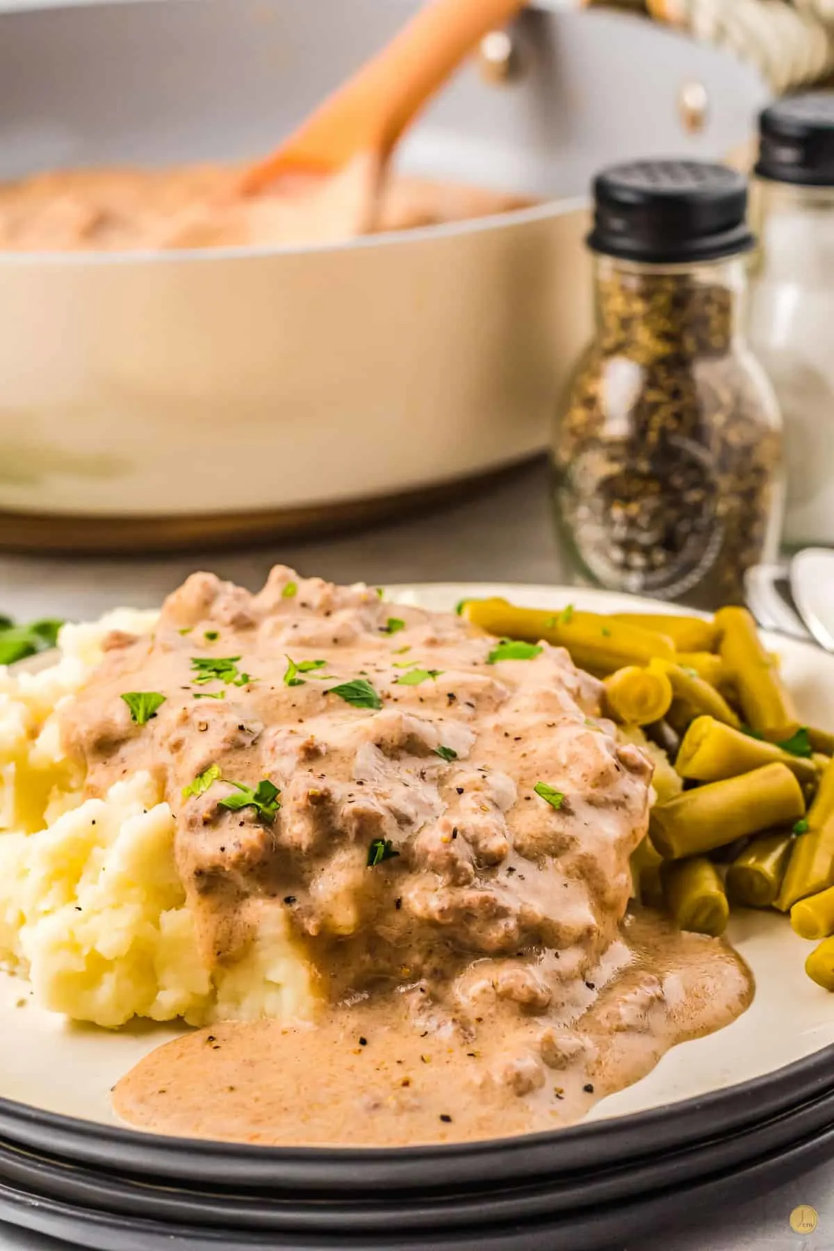 Close up of a white plate with a black rim filled with mashed potatoes topped with sausage gravy with a side of green beans.