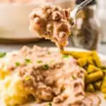 A white plate filled with mashed potatoes with sausage gravy with green beans on the side with a fork filled with sausage gravy being held in mid-air.