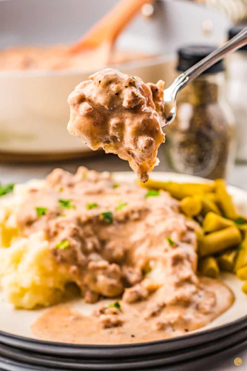 A white plate filled with mashed potatoes with sausage gravy with green beans on the side with a fork filled with sausage gravy being held in mid-air.