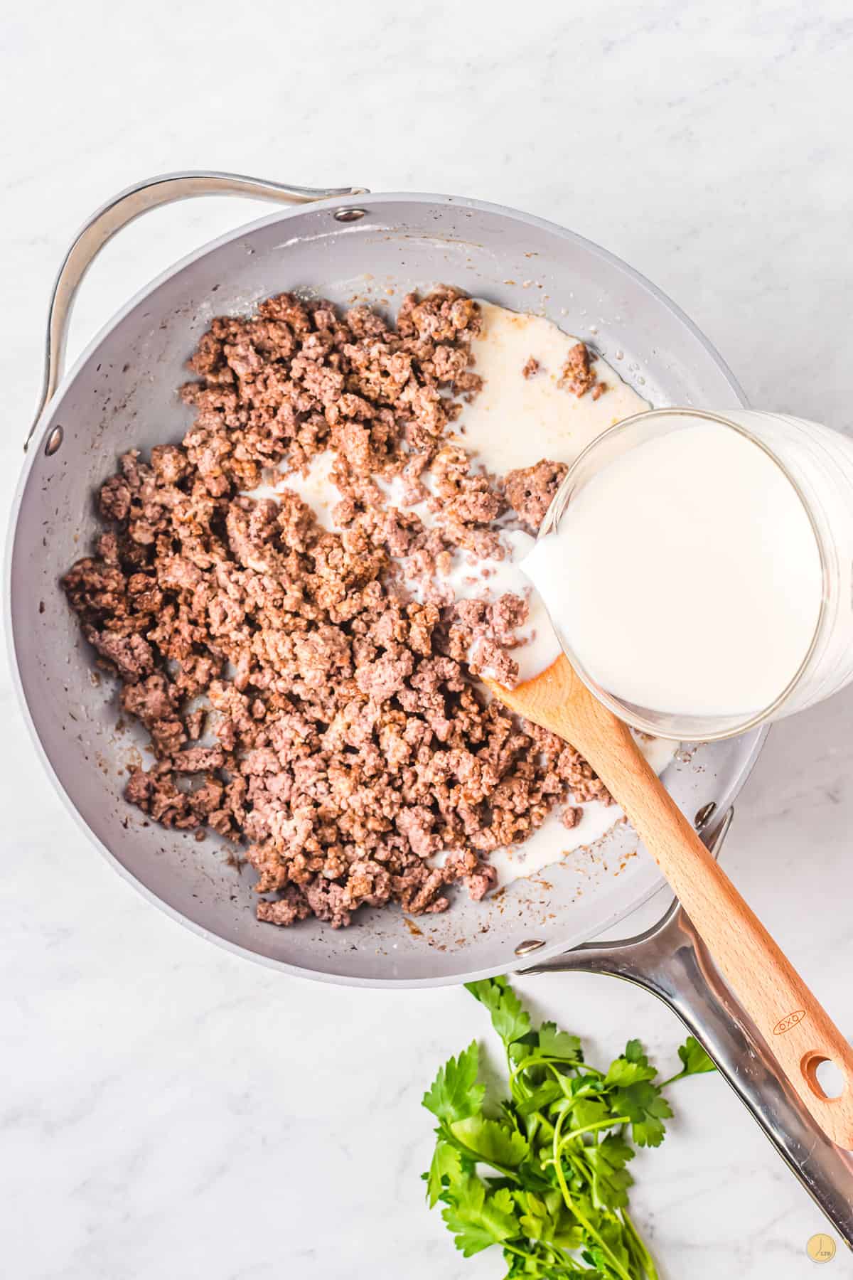 Top view of a pan with cooked mince and a wooden spoon in it with a jug of milk being poured in from the side. 
