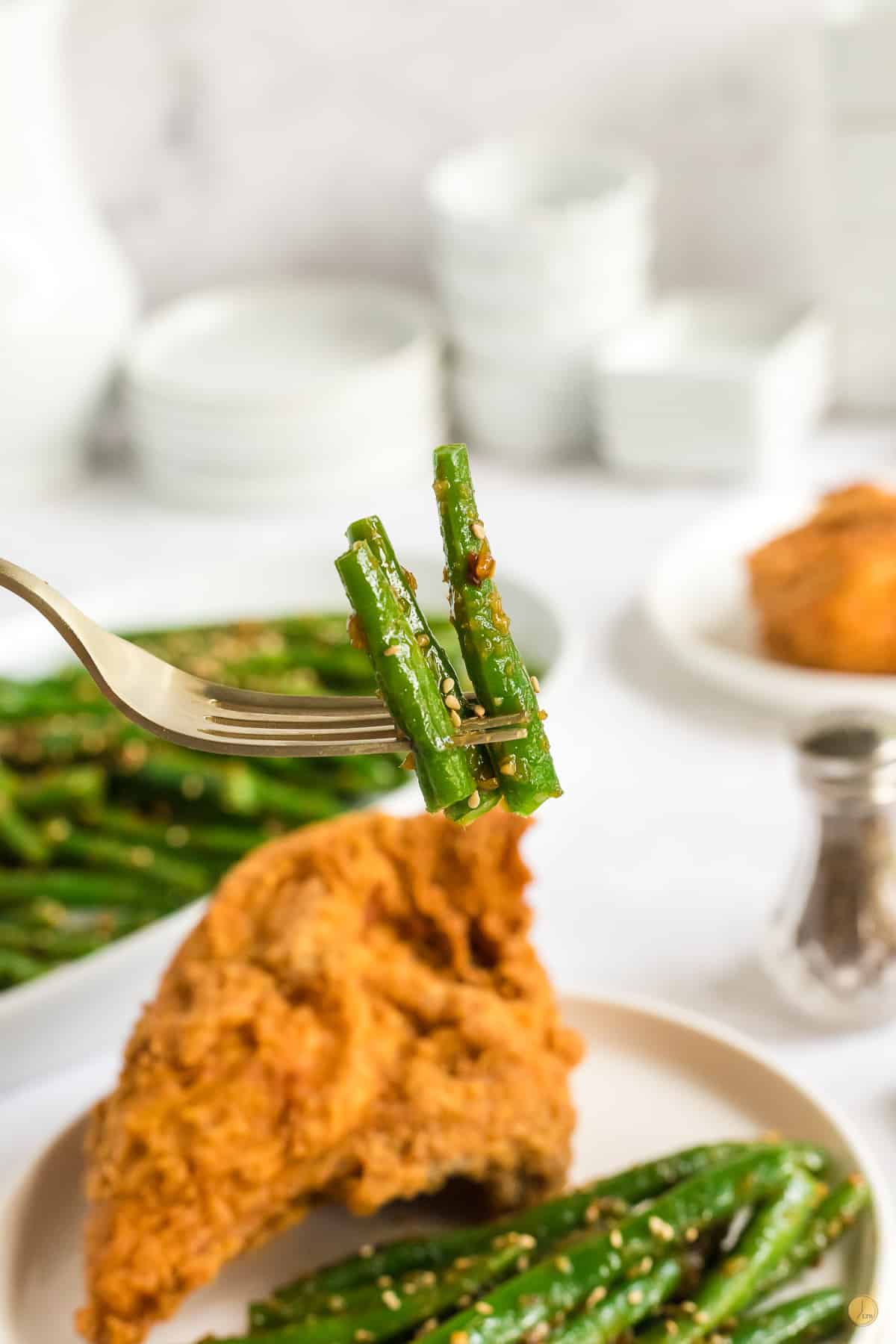 Close up of a fork in mid air holding 3 spicy green beans in the foreground, and a plate of crispy battered chicken on a plate with the rest of the spicy green beans. 