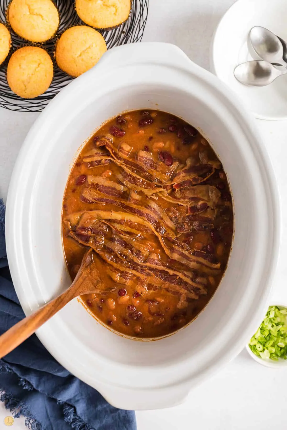 white slow cooker with beans, bacon, and a wood spoon.