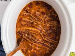 white slow cooker full of baked beans and a wood spoon