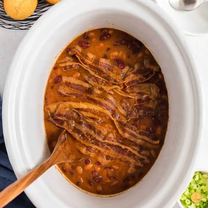 white slow cooker full of baked beans and a wood spoon