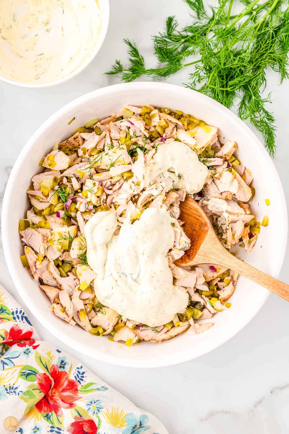 chicken salad in a bowl with a dollop of dressing being mixed in with a wooden spoon